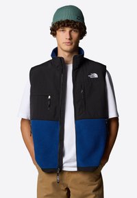 Czarny i niebieski polarowy kamizelka z wysokim kołnierzem, zamkiem z przodu i kieszeniami na zamek błyskawiczny. Noszona na białej koszuli, w komplecie z zieloną czapką z prążkowanej dzianiny.