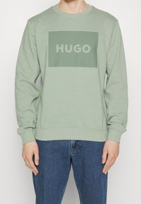 Sudadera de color verde claro hecha de tela suave, con un parche rectangular verde oliva que lleva el texto "HUGO" en blanco. Cuello redondo clásico.