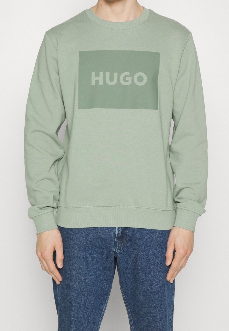 Sudadera de color verde claro hecha de tela suave, con un parche rectangular verde oliva que lleva el texto "HUGO" en blanco. Cuello redondo clásico.