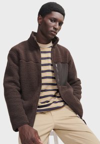 Brun fleecejakke med tekstureret stof, lynlås-lomme på brystet, høj krave, lagdelt over en stribet t-shirt og beige bukser.