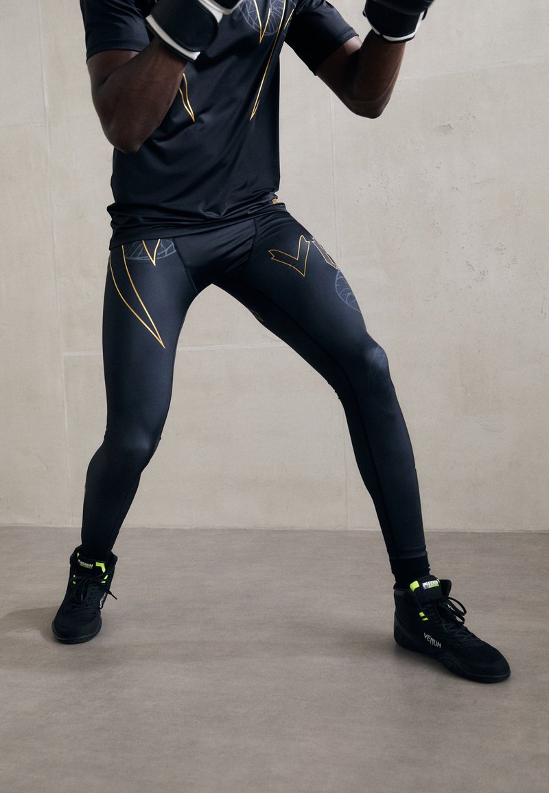 Schwarze Sportleggings mit gelben Akzentlinien und einem geometrischen Muster, kombiniert mit schwarzen Trainingsschuhen mit neongrünen Details.