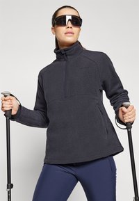 Pull noir en polaire avec un zip half-zip, poches latérales et détails de surpiqûre discrets. Paires de bâtons de ski tenus dans les mains.