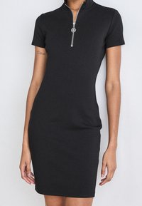 Czarna sukienka krótko rękawowa z wysokim kołnierzykiem i srebrnym suwakiem. Dopasowany fason z gładkiego, elastycznego materiału.