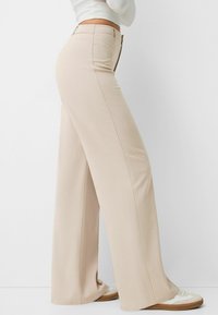 Beige vida byxor i ett mjukt tyg, med skräddarsydd passform, hög midja och synliga sömdetaljer, ihop med ljusa sneakers.