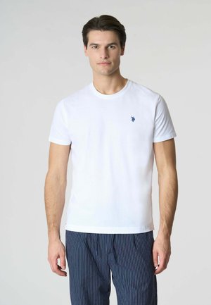 Uomo che indossa una semplice t-shirt bianca a maniche corte con un piccolo logo blu sul petto a sinistra e pantaloni blu scuro a righe sottili, in piedi contro uno sfondo chiaro.