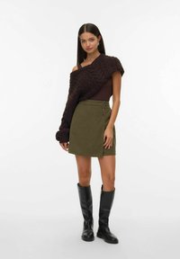 Ung kvinde iført en asymmetrisk brun kabelstrik sweater over den ene skulder, olivengrøn nederdel med knapper og sorte knæhøje støvler.