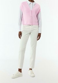 Jasnoróżowa dzianinowa kamizelka z dekoltem w serek i zapięciem na guziki, noszona na białej koszuli z kołnierzykiem, zestawiona z białymi dopasowanymi spodniami i trampkami.