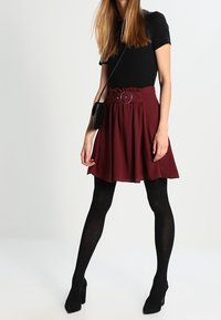 Frau mit einem schwarzen Kurzarmoberteil, einem bordeauxfarbenen Rock mit Gürtel, schwarzen Strumpfhosen, schwarzen High Heels und einer kleinen schwarzen Umhängetasche.
