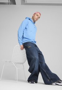 Sudadera con capucha de color azul claro con un logo, combinada con unos vaqueros oscuros, holgados y desgastados. La persona se apoya en una silla blanca de alambre.