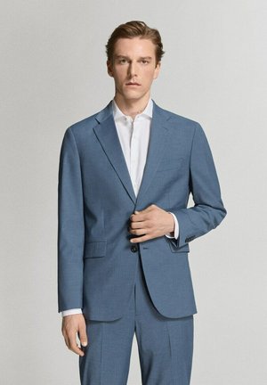 Joven con chaqueta de traje azul claro y pantalones a juego sobre una camisa blanca, posando frente a un fondo claro y liso.