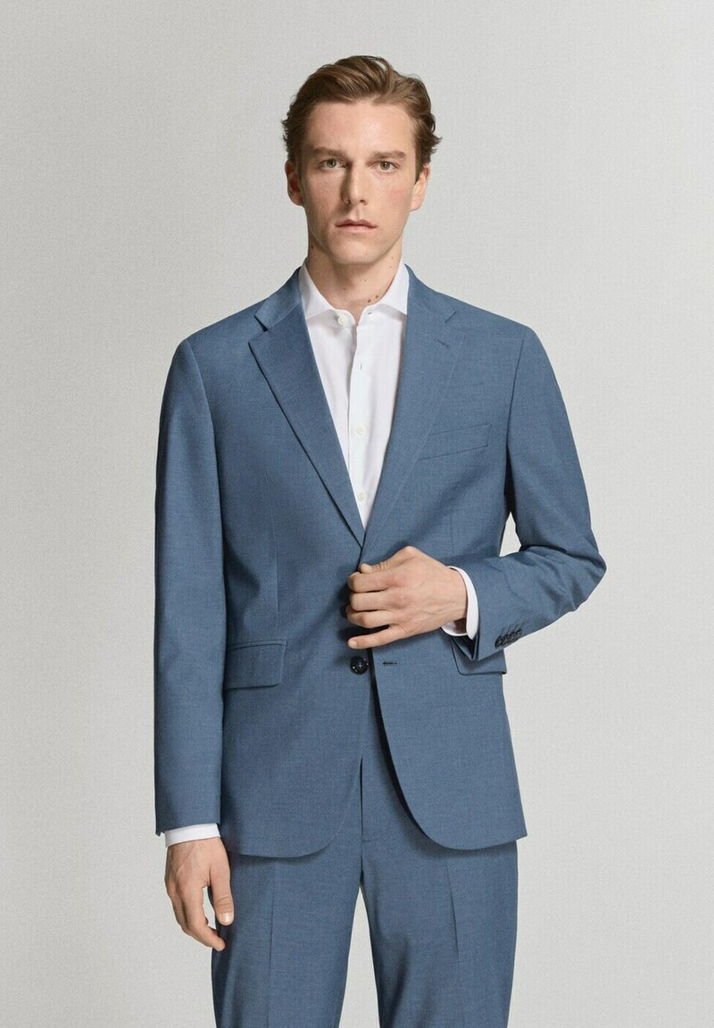 Joven con chaqueta de traje azul claro y pantalones a juego sobre una camisa blanca, posando frente a un fondo claro y liso.