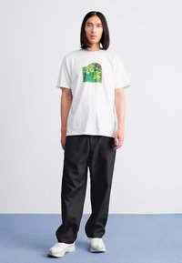 White t-shirt featuring a colorful graphic on the chest, paired with black loose-fitting pants and white sneakers, on a simple background.