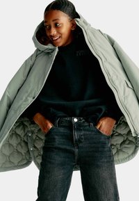 Giacca esterna puffosa di un grigio chiaro, aperta, che rivela un rivestimento interno trapuntato. Felpa nera sotto e jeans di denim scuro con tasche anteriori.