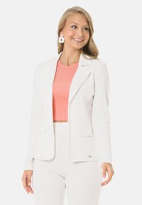 Weißer gerippter Blazer mit Revers, vorderen Taschen und einem silbernen Herz-Akzent, über einem korallenfarbenen gerippten Crop-Top getragen.
