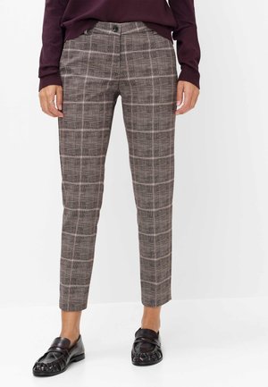 Kvinde iført tætsiddende brune ternede bukser, mørkerød sweater og sorte loafers står på hvid baggrund.
