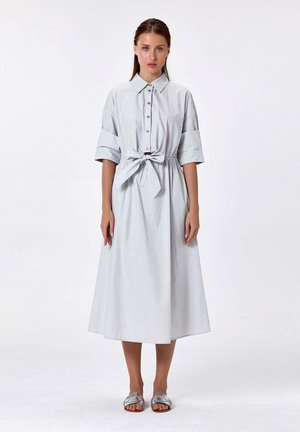 Femme debout portant une robe-chemise midi gris clair avec manches retroussées, boutons devant, ceinture nouée à la taille et sandales argentées à enfiler.