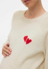 Persona embarazada con un suéter crema y un diseño de corazón roto rojo, sosteniendo suavemente su barriga contra un fondo blanco liso.