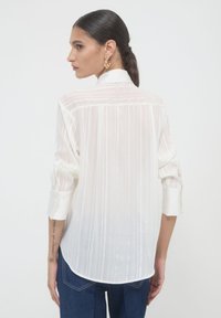 Donna con capelli scuri raccolti in una coda di cavallo, che indossa orecchini a cerchio dorati, una blusa bianca trasparente a maniche lunghe e jeans blu, di spalle.