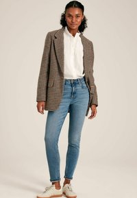 Blazer marrón y crema con cuadros sobre una blusa blanca, combinado con jeans ajustados azul claro y zapatillas crema. Presenta solapas clásicas.