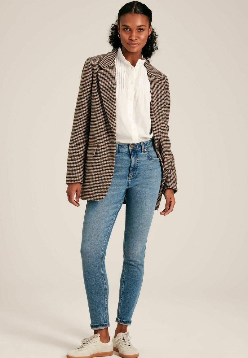 Blazer marrón y crema con cuadros sobre una blusa blanca, combinado con jeans ajustados azul claro y zapatillas crema. Presenta solapas clásicas.