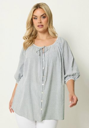 Blonde vrouw die een losse, gestreepte blouse draagt met knopen en een strik bij de halslijn, gecombineerd met een witte broek, staand tegen een effen achtergrond.