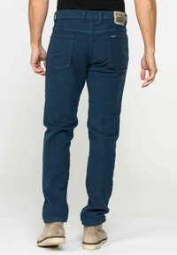 Pantaloni in cotone blu navy con gamba dritta, tasche posteriori e una toppa in pelle con logo sulla vita. Si abbinano a calzature beige.
