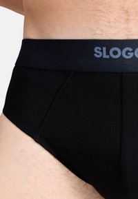 Musta puuvillatrimmeri, jossa on sileä, joustava vyötärö, johon on kirjattu "SLOGAN" harmaalla. Ominaisuuksiin kuuluu muotoiltu malli ja teksturoitu kangas.