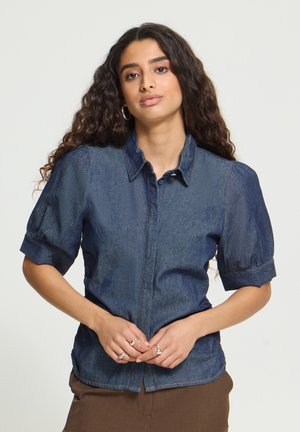 Ung kvinde med langt krøllet hår iført en kortærmet denimskjorte og brune bukser, stående med hænderne foldet foran.