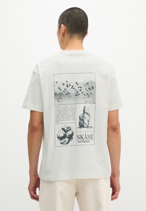 Man draagt wit t-shirt met zwarte print van vliegende vogels, rotsen en tekst over Skåne wetlands op de rug, tegen een effen lichte achtergrond.