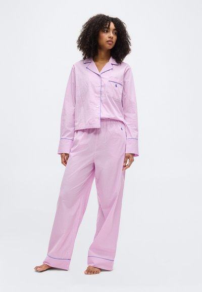 Γυναίκα που στέκεται ξυπόλητη, φορώντας ένα χαλαρό ροζ ριγέ σετ πυτζάμας με μακριά μανίκια, πουκάμισο με κουμπιά και φαρδιά παντελόνια, μπροστά σε ένα απλό φόντο.