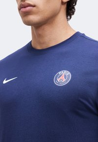 Άνδρας που φορά μπλούζα Paris Saint-Germain σε σκούρο μπλε χρώμα με το λογότυπο του συλλόγου και λευκό λογότυπο Nike στο στήθος, περικομμένη στον λαιμό και στους ώμους.