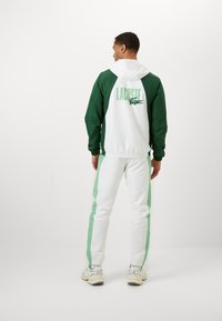 Vīriešu Lacoste sporta uzvalks, kas izceļas ar balto un zaļo krāsu kombināciju. Jakai ir kapuce, zaļas piedurknes un logo uz muguras. Biksēm ir saskaņoti sānu paneļi.