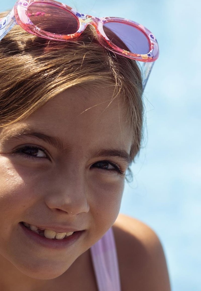 Girl with long brown hair wearing pink and clear sunglasses, looking directly at the camera. Soft focus background with blue water.