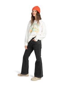 White sweatshirt with a graphic print, black flared jeans, and a bright orange knit beanie. Beige footwear completes the outfit.