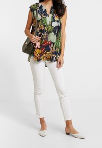 Blusa floreale di colore blu navy con motivi verde e giallo, pantaloni bianchi slim-fit e mule bianche perforate. Borsa verde oliva.