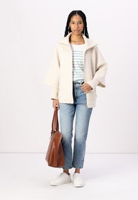 Beige oversized jakke med brede ærmer over en lyseblå og hvidstribet skjorte, parret med lys vaskede jeans og en brun tote bag.