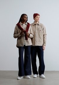 Két modell visel egyező bézs színű, steppelt ingeket logós díszítéssel, amelyeket sötét, bővülő szárú farmerrel és fehér cipővel párosítanak. Az egyik modellen piros sál van.