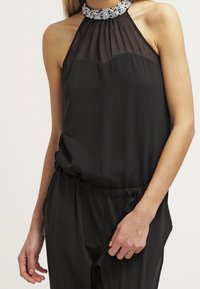 Top halter negro sin mangas con yugo transparente y cuello alto decorado, combinado con pantalones negros a juego.