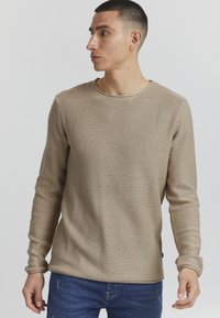 Beige strukturierte Langarmbluse mit Rundhalsausschnitt und entspannter Passform, kombiniert mit blauen Jeans.auffälliges Rippmuster über die gesamte Oberfläche.