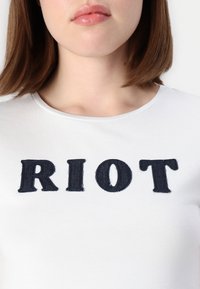 Bela bomba majica iz bombaža, na kateri so v velikih mornarsko modrih vezenih črkah napisana beseda "RIOT," z klasičnim okroglim izrezom in gladko teksturo tkanine.