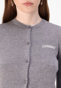 Grå strikket cardigan med rund hals, fem mørke knapper, og teksten "2ND DAY" brodert i lys grå på venstre side.