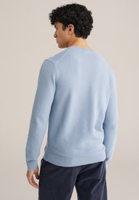 Svetlo modra teksturirana majica s rebrastim izrezom na vratu in manšetami, z dolgimi rokavi in sproščenim krojem. Nosi se s temnimi hlačami.