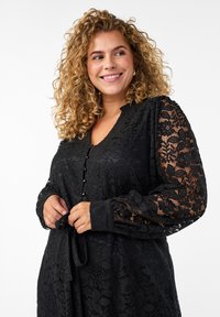 Čierne čipkované šaty s dlhými rukávmi, véčkovým výstrihom a gombíkovými detailmi. Látka má kvetinový vzor s textúrovaným povrchom a viazací opasok.