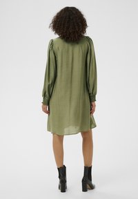 Robe vert olive à manches bouffantes avec poignets froncés, dotée d'une coupe décontractée et d'un tissu léger. Portée avec des bottines noires.