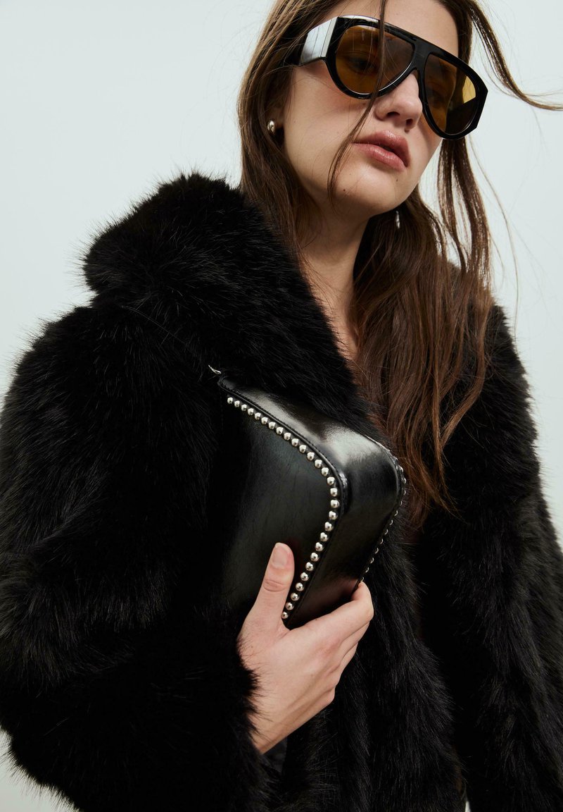 Cappotto in pelliccia sintetica nera con colletto, tenuto in mano con una mano che stringe una borsa di pelle nera decorata con borchie argentate. Indossati occhiali da sole neri di grandi dimensioni.