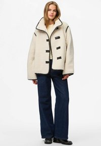 Chaqueta extragrande de piel sintética tipo borrego en color crema con ribete y botones negros, con cuello alto, combinada con vaqueros anchos de pierna en azul oscuro.