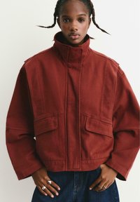 Rode cropped jas van stevig materiaal, met een hoge kraag, grote voorkruiken en brede mouwen, gedragen met donkerblauwe jeans.