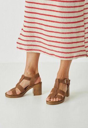 Brown woven high-heel sandals with an open toe, featuring cross straps and a buckle closure, mounted on a wooden textured heel.