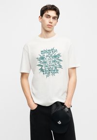 Νέος άνδρας ντυμένος με λευκό T-shirt με το γράφημα "Earth Tripper" και μαύρο σορτς, κρατώντας ένα μαύρο καπέλο από δίχτυ σε ένα χέρι.