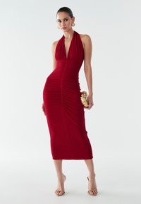 Raudona halterio suknele su raukšlėmis, prigludusi siluetas ir iki kelių ilgio. Modelis laiko auksinę rankinę ir užsima neutralias aukštakulnes batus.